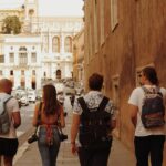 Four tourists with backpacks walking on a street in Rome, Italy, with historic architecture around.